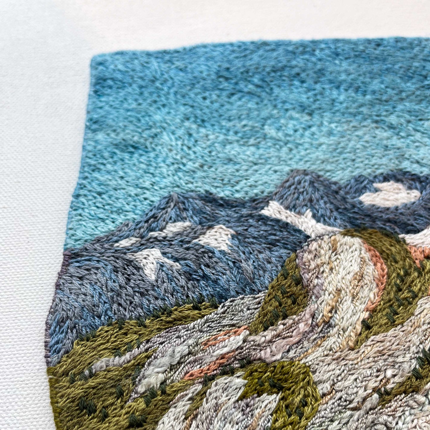 Close-up of embroidered wall art: mountain peaks with snow details, textured slopes, and detailed stitching.