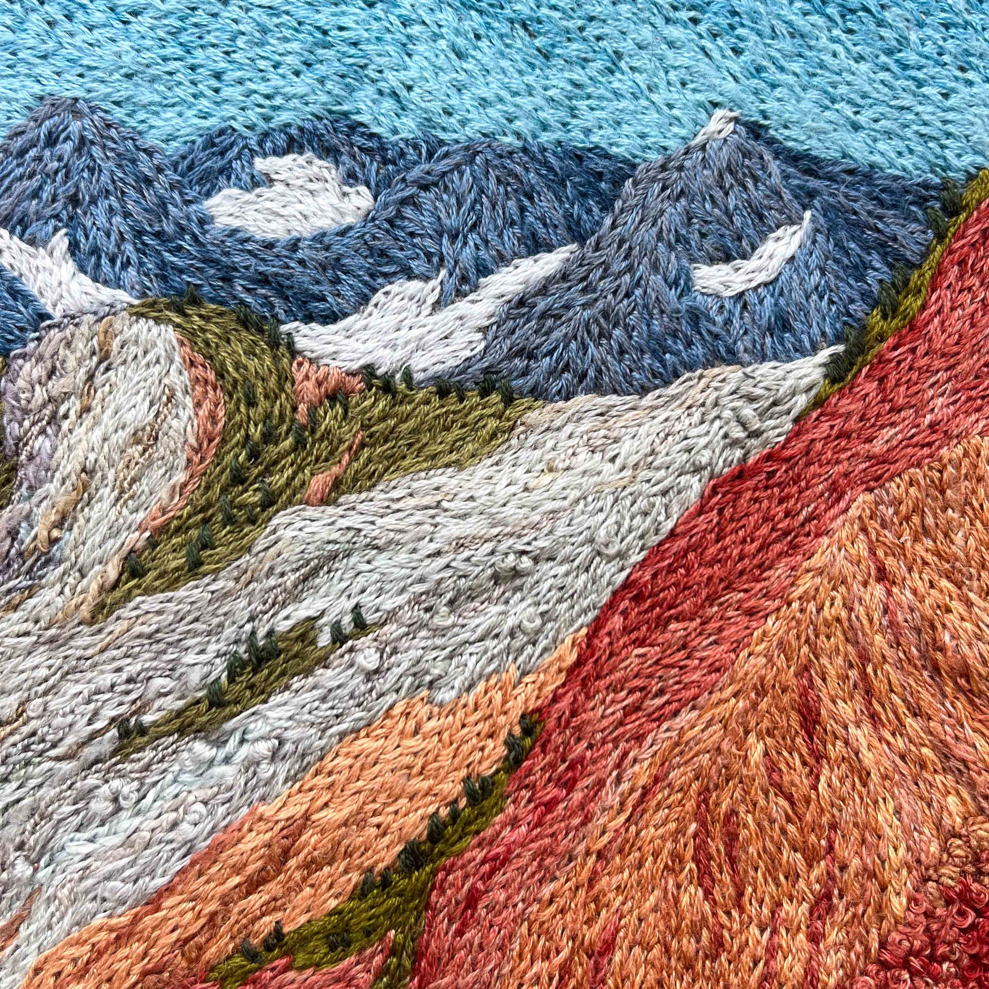 Close-up of embroidered wall art: mountain peaks with snow details, textured slopes, and vibrant stitching.