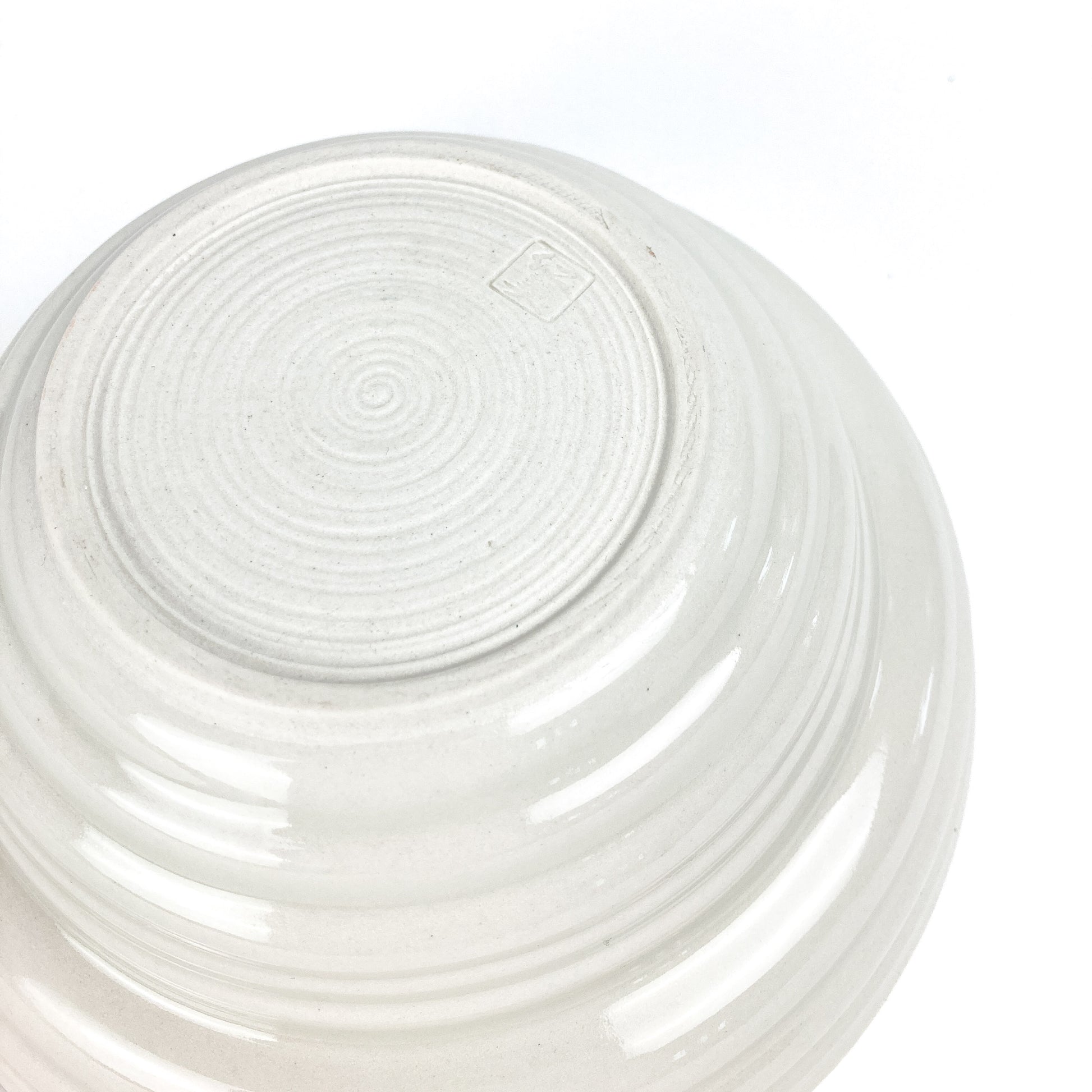 Bottom view of a round ceramic bowl showing concentric circular markings and a small, square maker's mark. The bowl has textured horizontal ridges.
