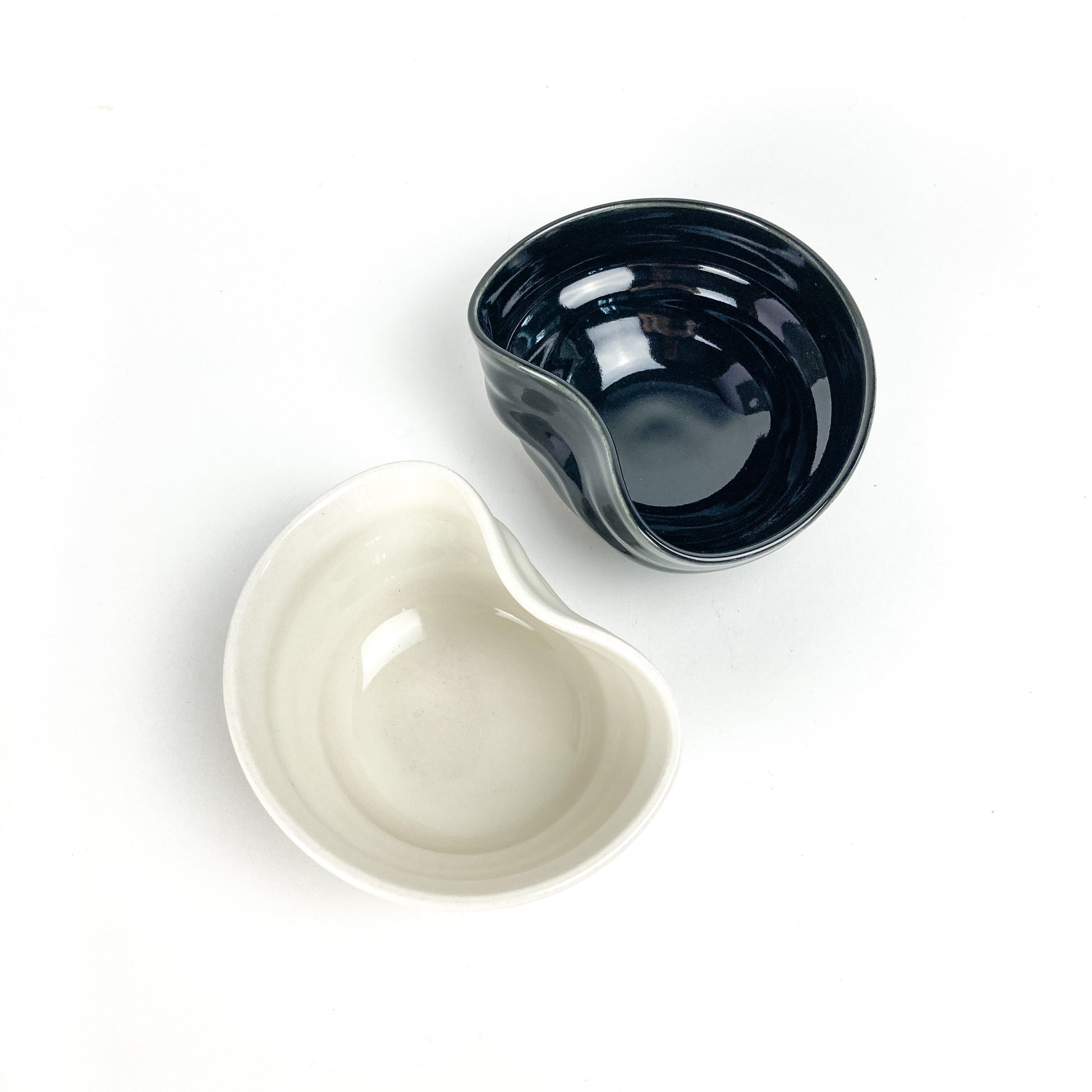 Two small, concave ceramic bowls: one dark and glossy, the other light with a matte finish, both with an organic, asymmetrical shape.