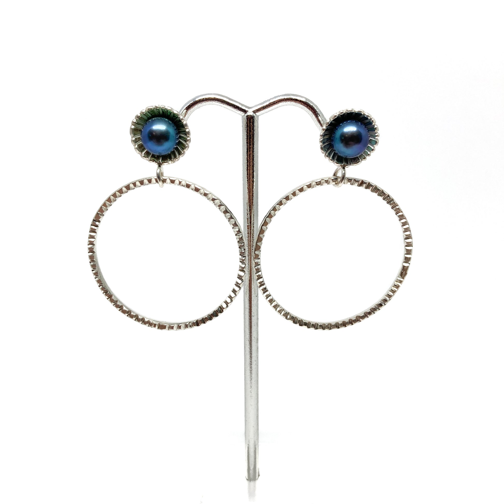 A pair of silver hoop earrings on a stand. Each earring features a dark, iridescent pearl set above a textured hoop.