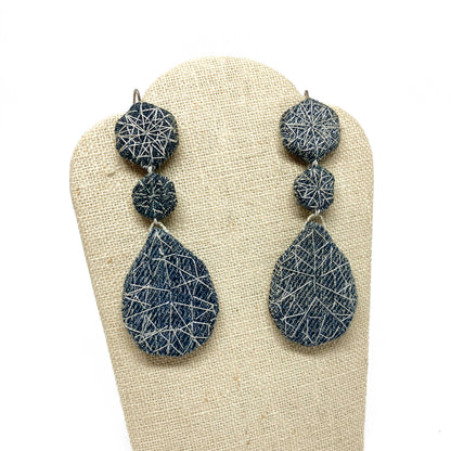 "Patched Coronation (Street)" earrings by Louise Perrone displayed on a textured stand. Each earring features three textured, fabric-patched shapes.