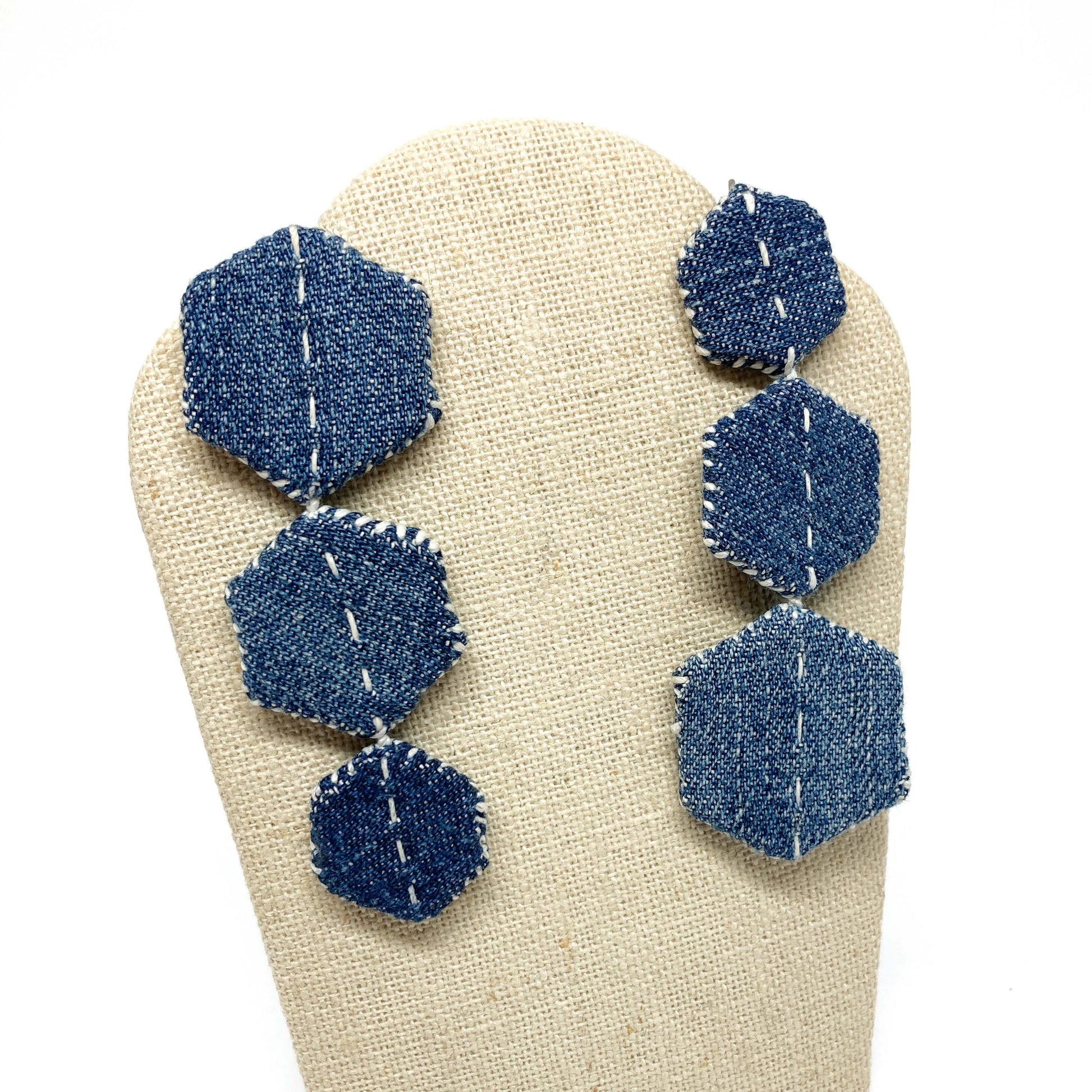 "Patched Trio" earrings by Louise Perrone displayed on a textured stand. Each earring features three denim hexagons with white stitching.