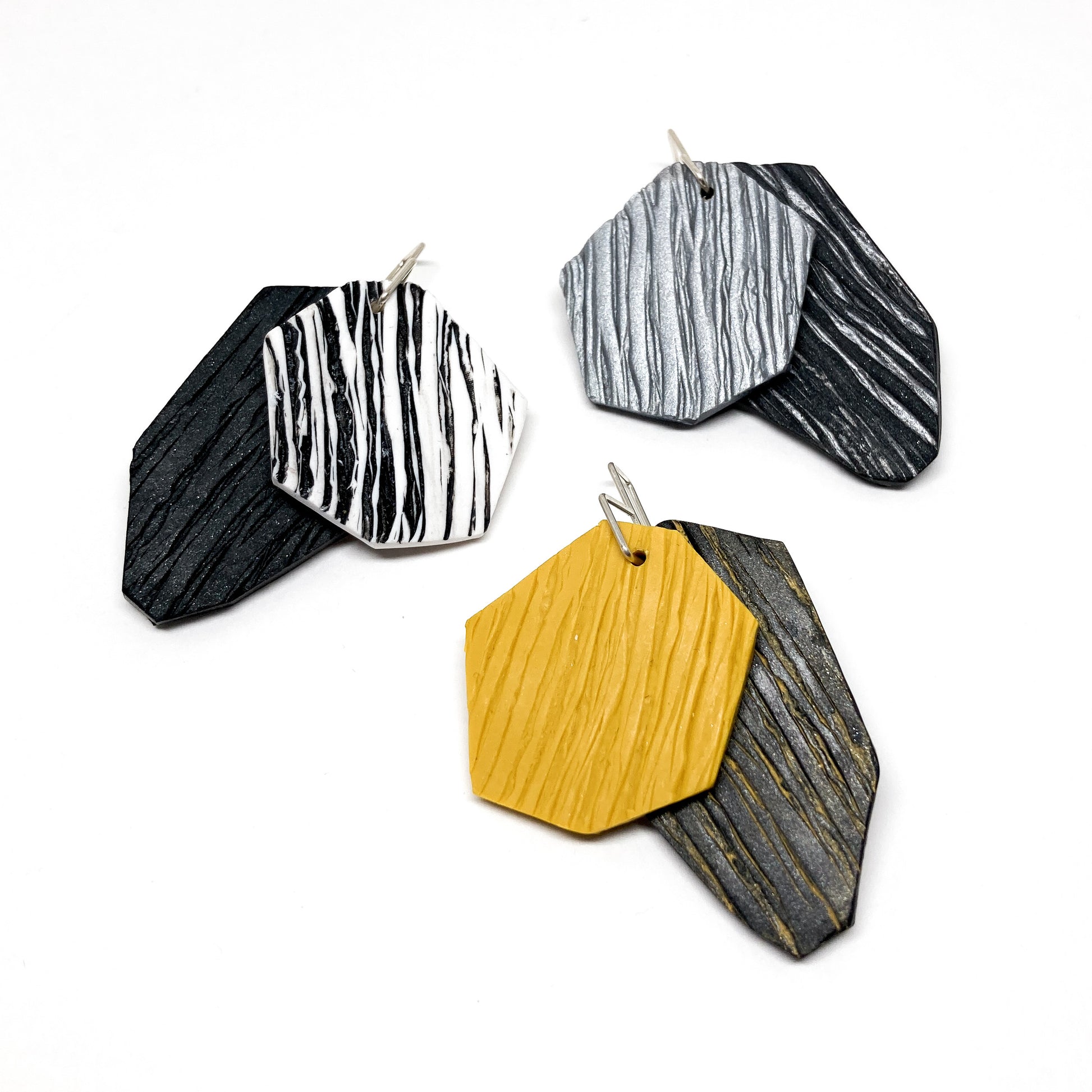 Three pairs of textured, geometric earrings: black with white stripes, silver with black stripes, and yellow with black. They hang from silver hooks.
