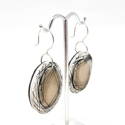 Side view of round dangle earrings with abstract black line design on a layered circular base, hanging on a metal stand.