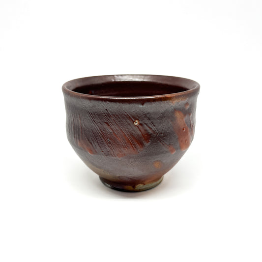 A textured, wood-fired ceramic tea bowl features diagonal markings and variations in tone.