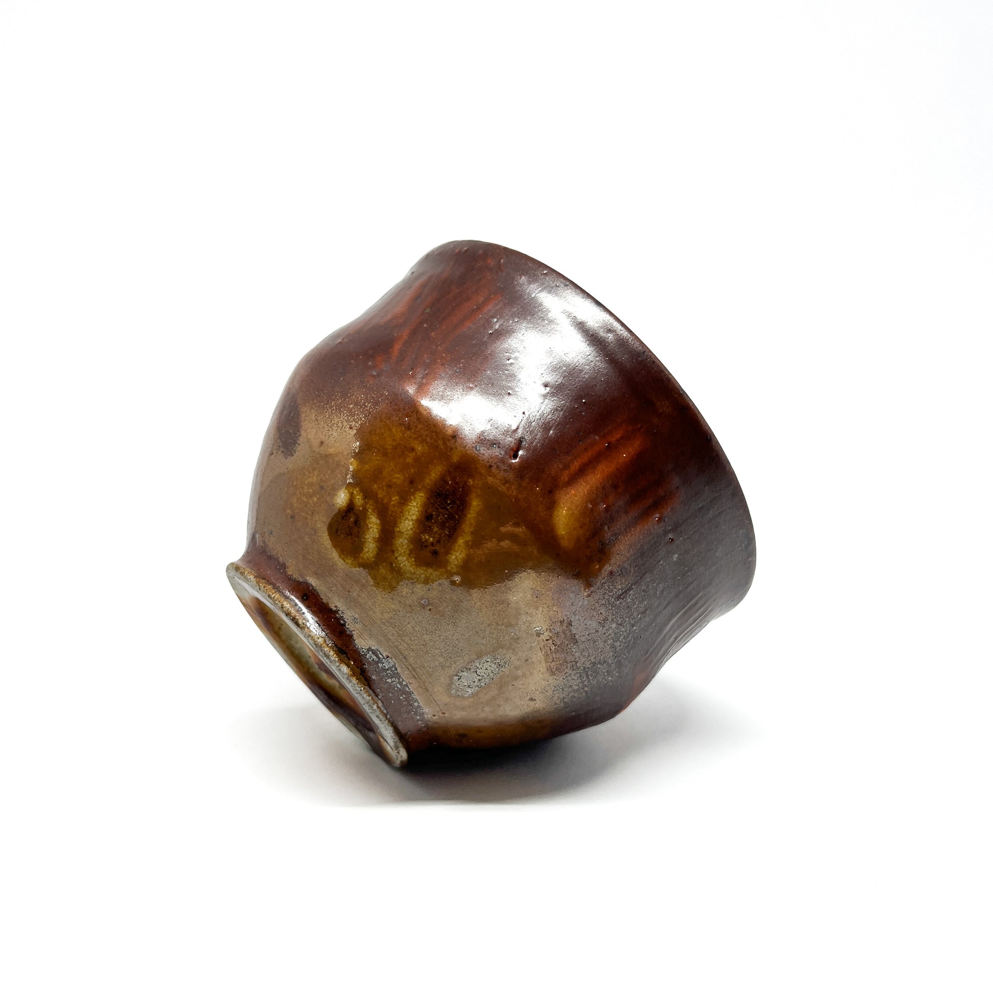 Angled view of a handcrafted, dark, glossy tea bowl with earthy tones and a slightly uneven rim.