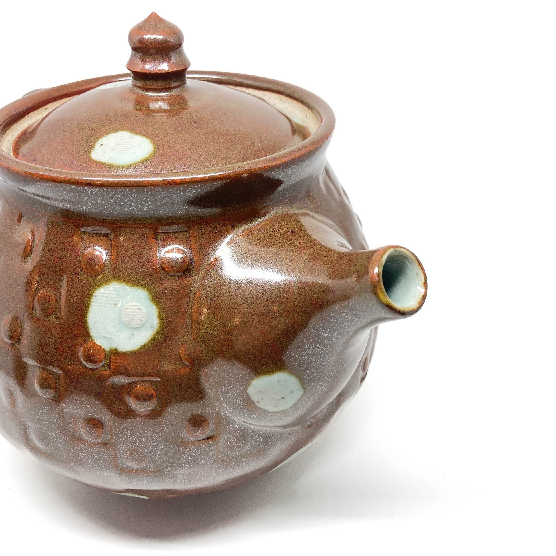 Close-up of a brown ceramic teapot with a lid and spout, featuring textured details and light-colored circular designs.