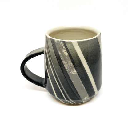 A ceramic mug, possibly wood-fired, with diagonal stripes in shades of gray and white, and a dark handle. The inside is a light tone.