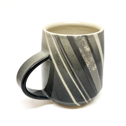 A wood-fired ceramic mug with diagonal stripes in shades of gray and white, and a dark handle. The mug's interior is a light color.