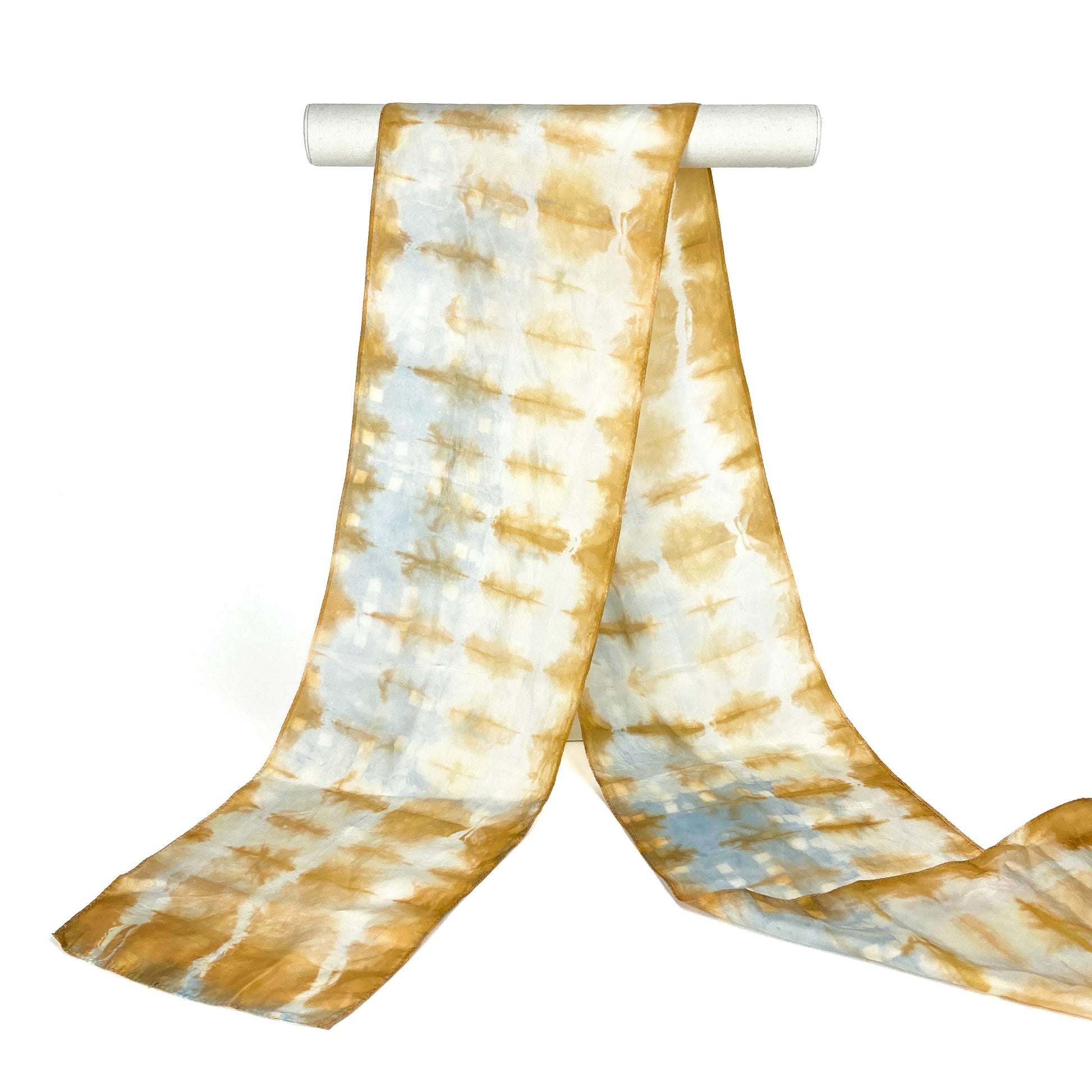 A cutch tree bark silk scarf with a horizontally textured shibori tie-dye pattern hangs from a white cylindrical bar.