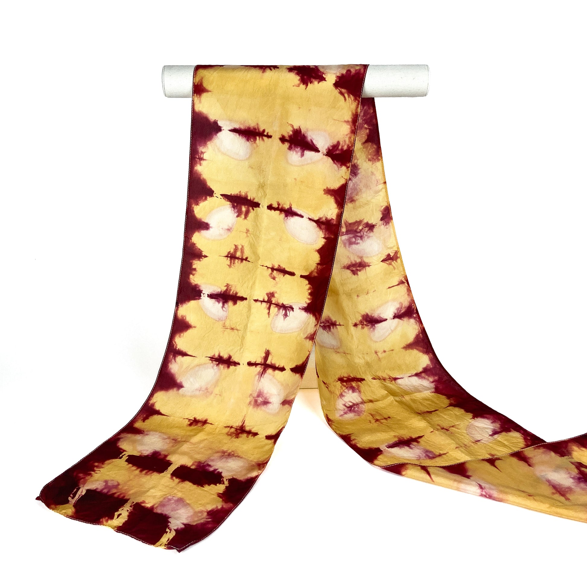A Rhubarb Lac silk scarf with a distinctive shibori tie-dye pattern hangs gracefully from a white cylindrical bar.