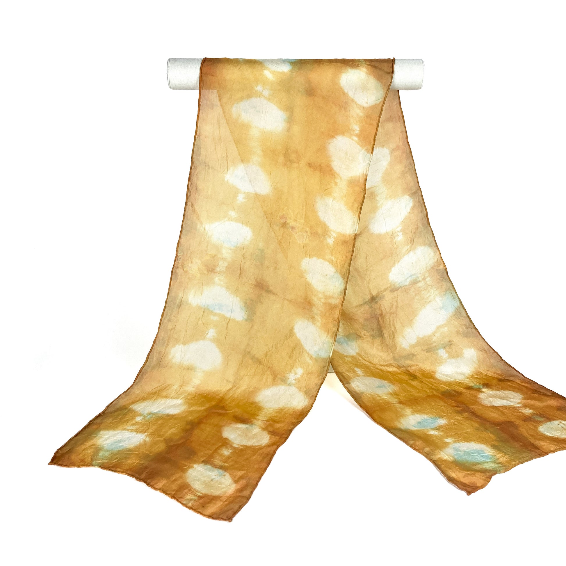 A Fustic silk scarf with a unique shibori tie-dye pattern featuring resist circles hangs from a white cylindrical bar.