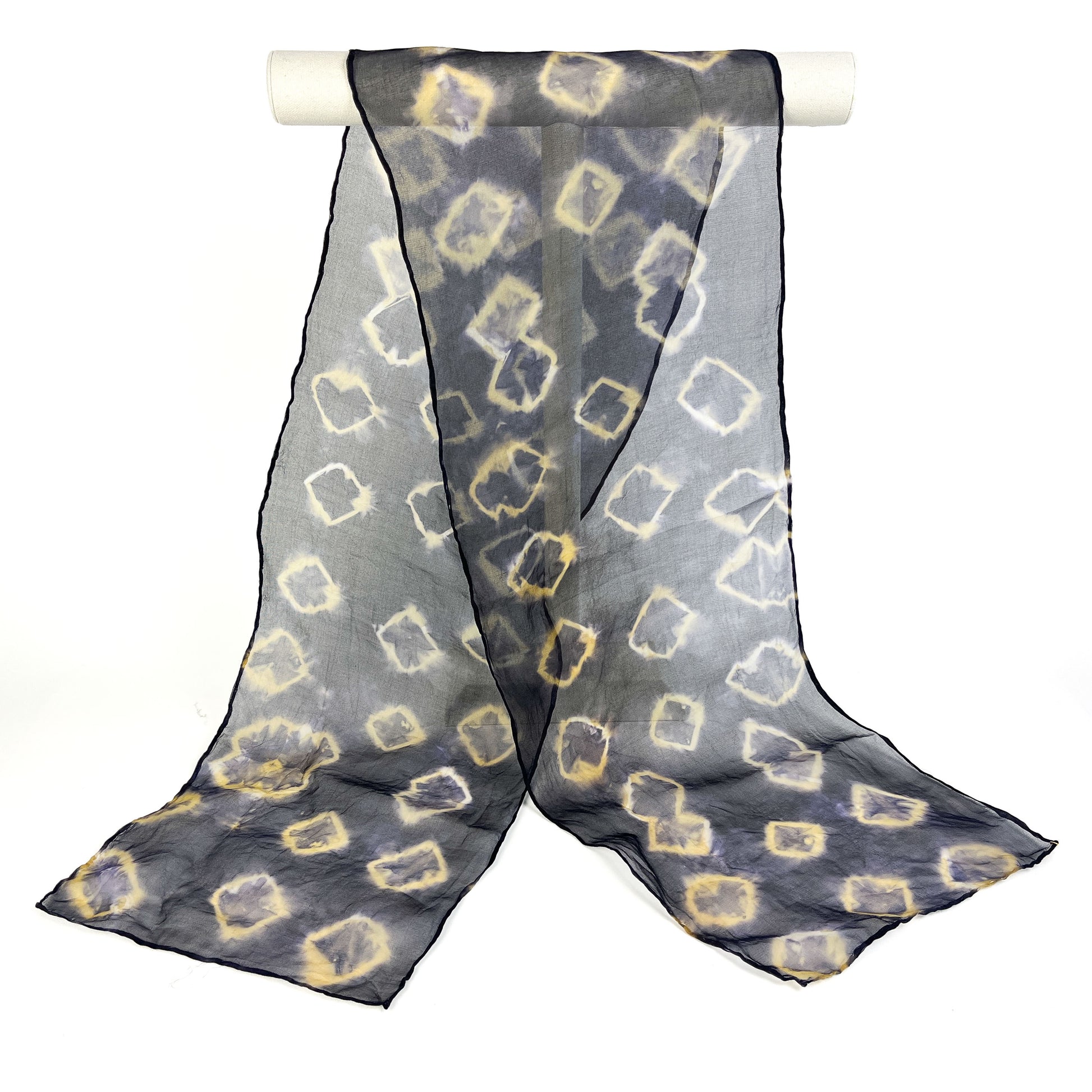 A Black Bean Hull silk scarf with a resist shibori pattern of outlined squares hangs from a white cylindrical bar.