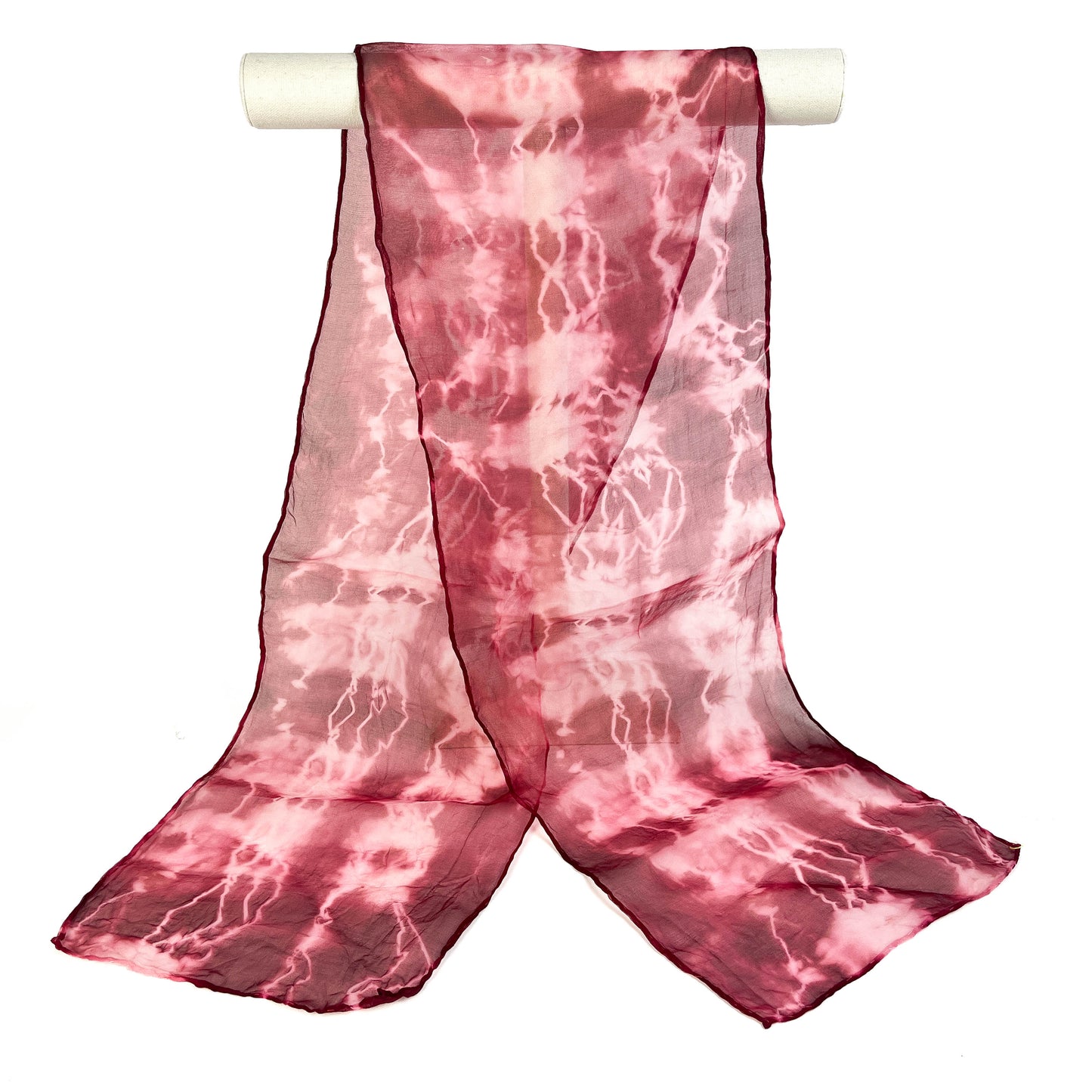 A pink shibori hand-painted pure silk scarf with a crackled pattern drapes elegantly from a white cylindrical bar.