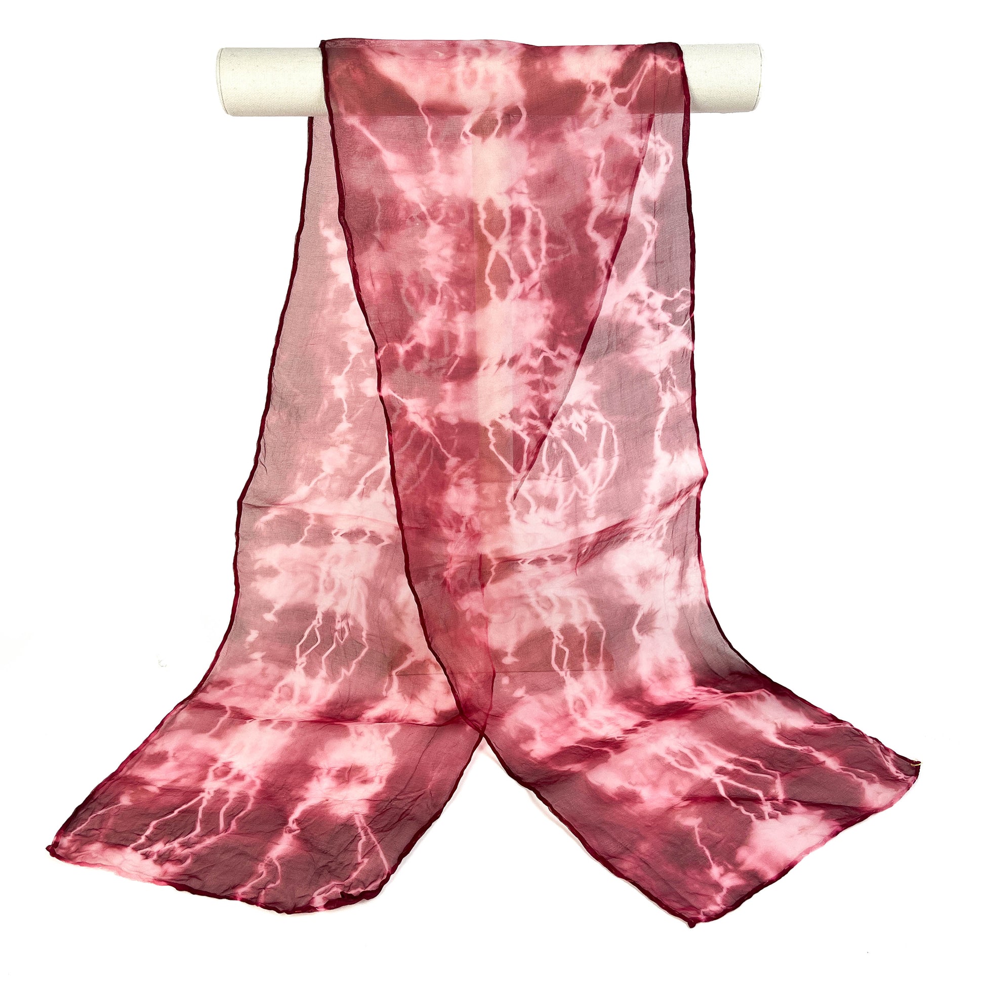 A pink shibori hand-painted pure silk scarf with a crackled pattern drapes elegantly from a white cylindrical bar.