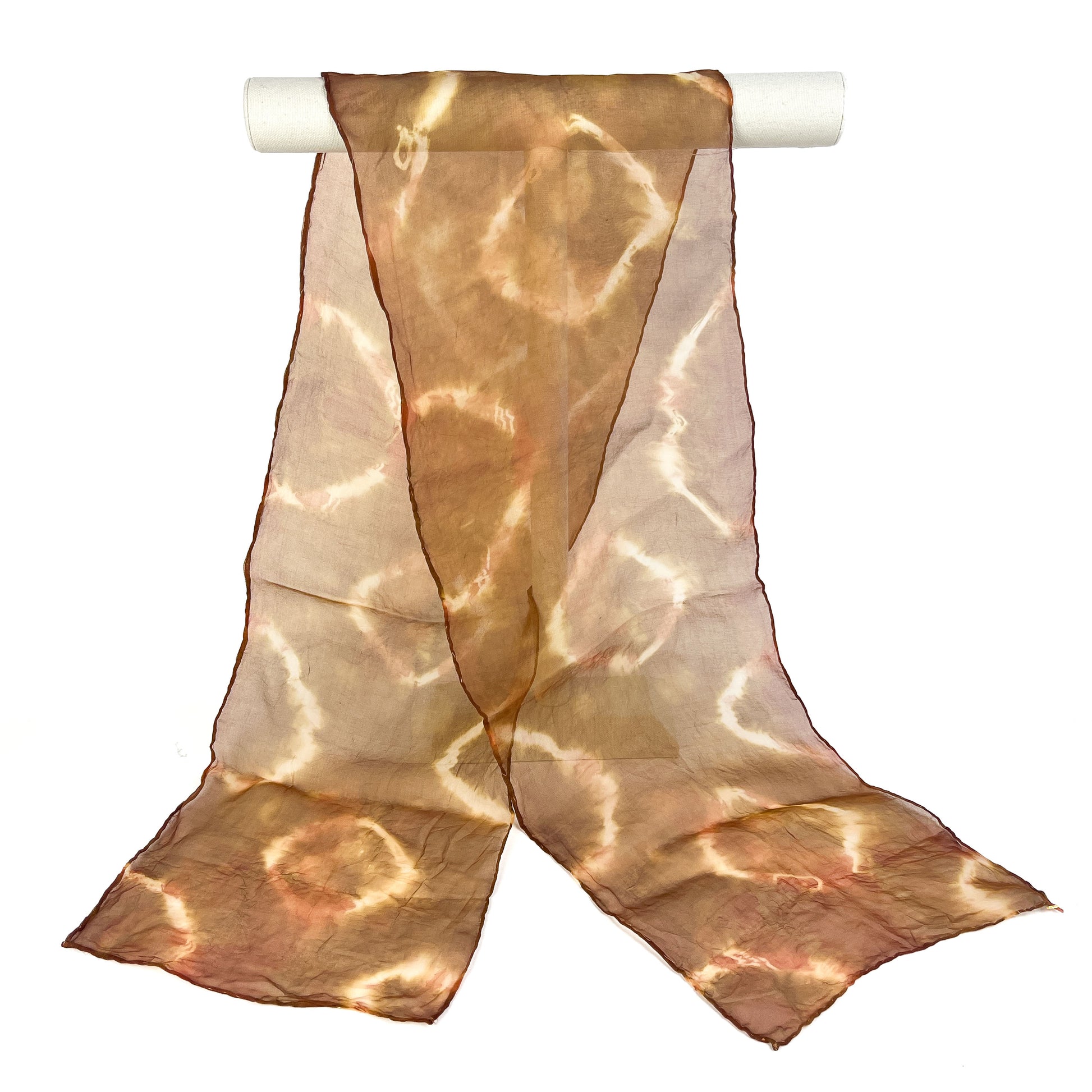 A Tea silk scarf with a circular resist shibori tie-dye pattern hangs softly from a white cylindrical bar.