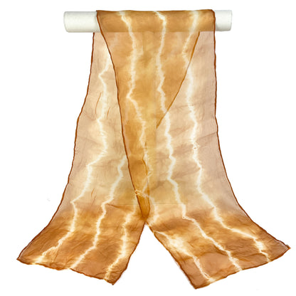 A Walnut Hull silk scarf with a wavy, horizontal shibori tie-dye pattern drapes from a white cylindrical bar.