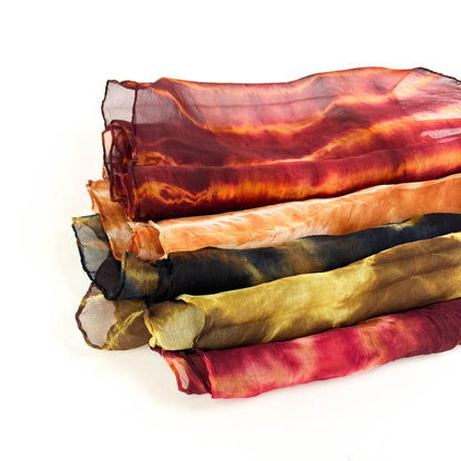 A neatly stacked pile of six sheer silk scarves showcases a variety of vibrant, blended tie-dye patterns and textures.
