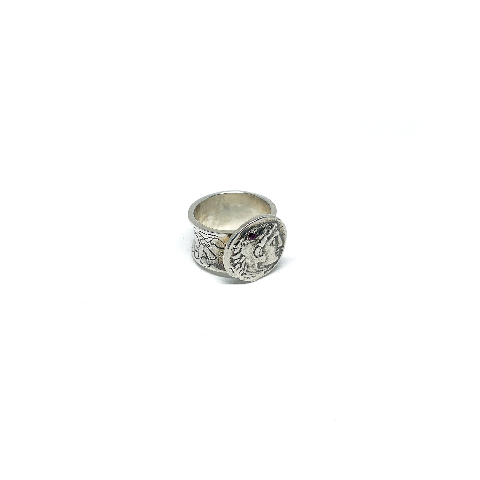 A silver ring with a textured band and an attached antique coin featuring a profile and a small red stone.