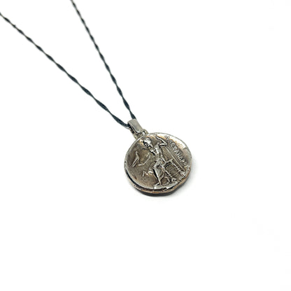 Close-up of a silver necklace with a dark chain and a round pendant featuring an engraved figure and Greek inscription.