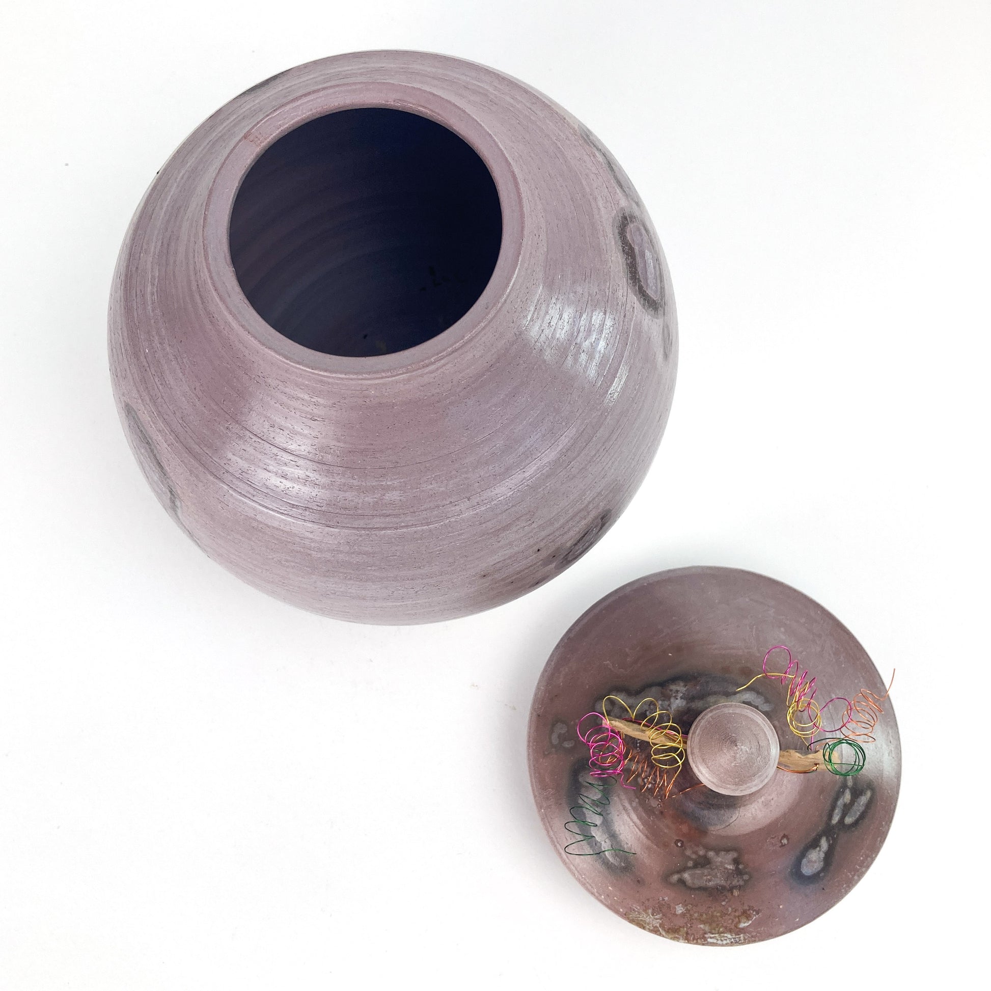 Top-down view of a lavender ceramic jar with its lid slightly askew, revealing the dark interior. Colorful wire on the lid.