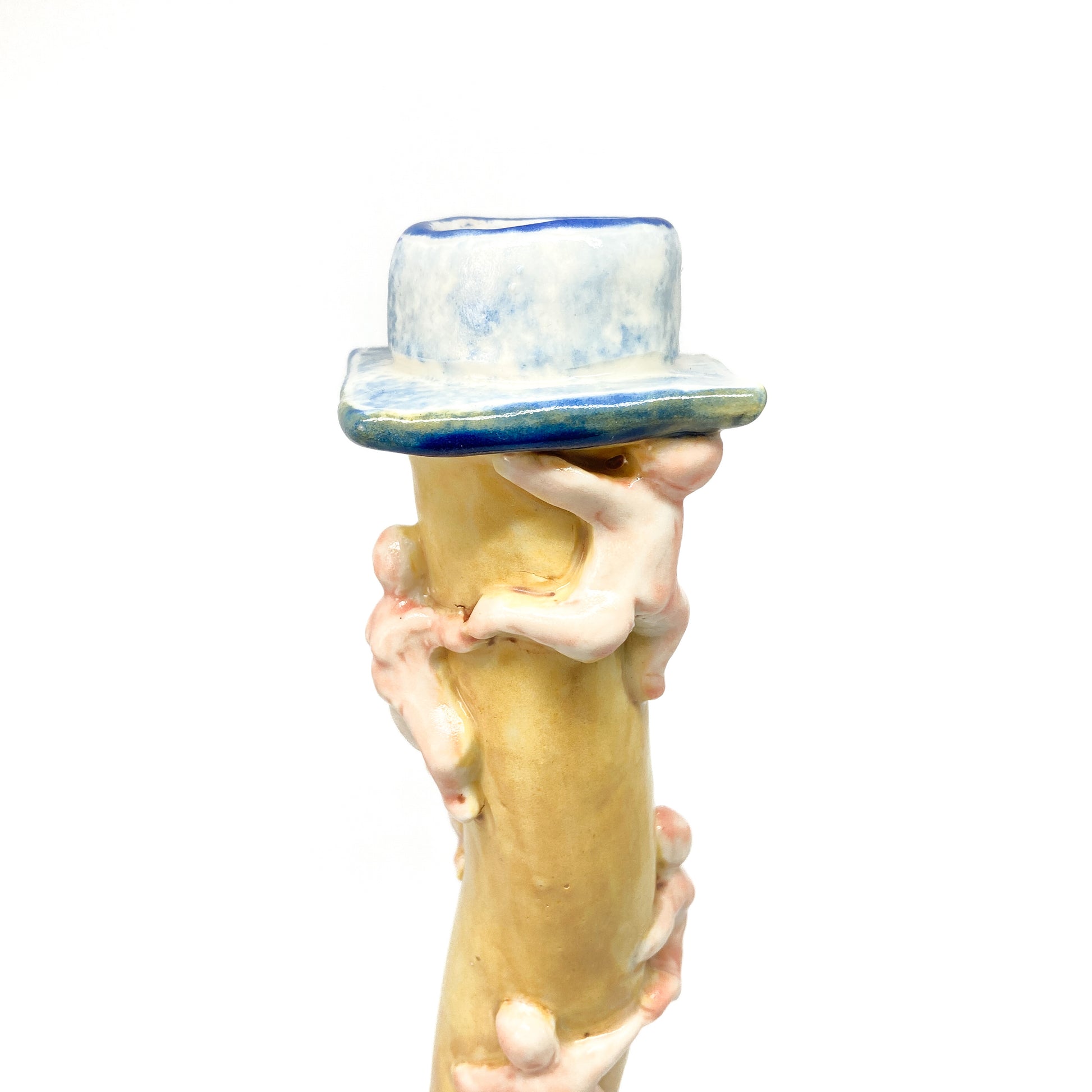 Top portion of a yellow ceramic candlestick with small, light-colored figures appearing to climb the wavy stem towards the blue and white top.