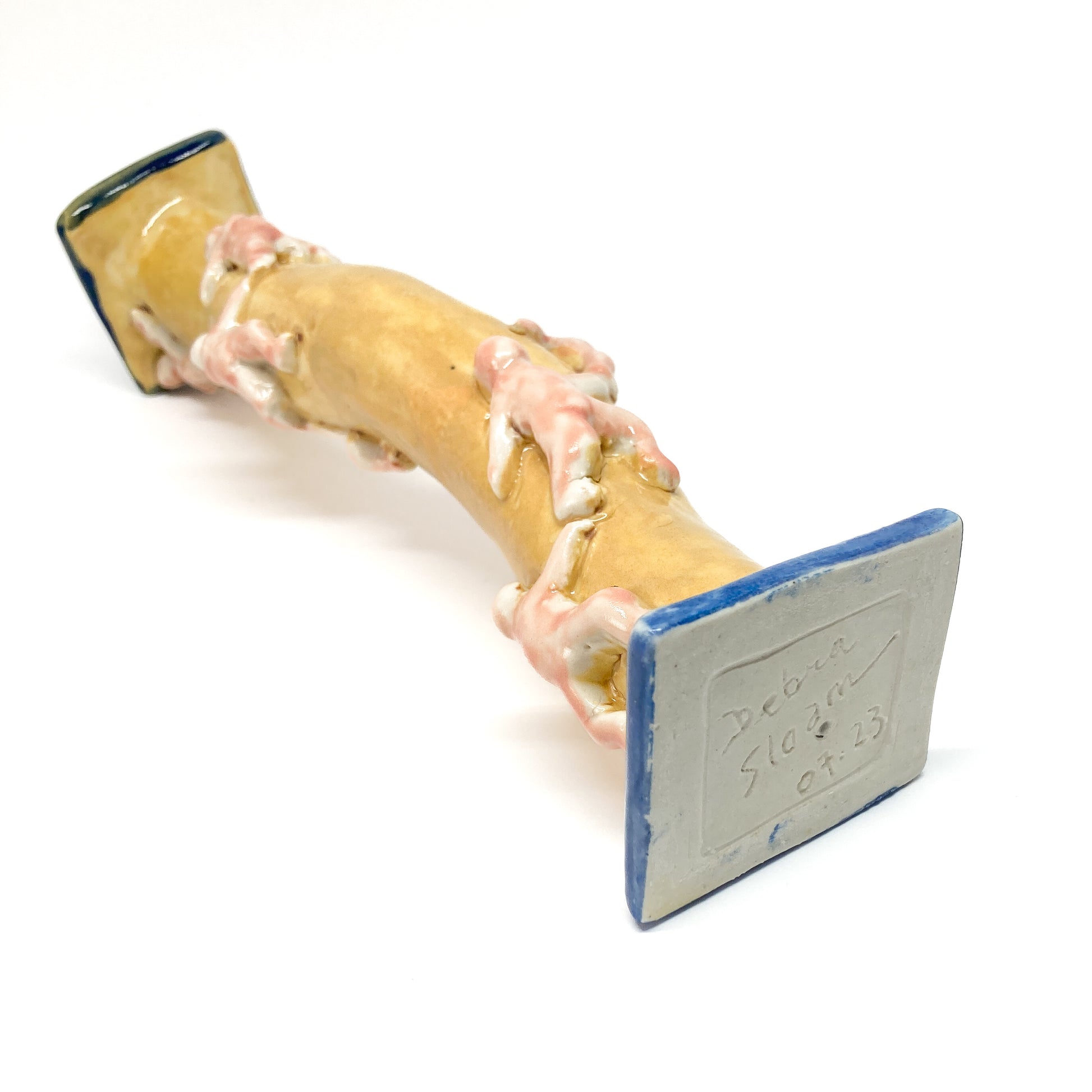 A yellow ceramic candlestick with small, light-colored figures climbing its wavy form lies horizontally. The base has blue trim and an inscription.