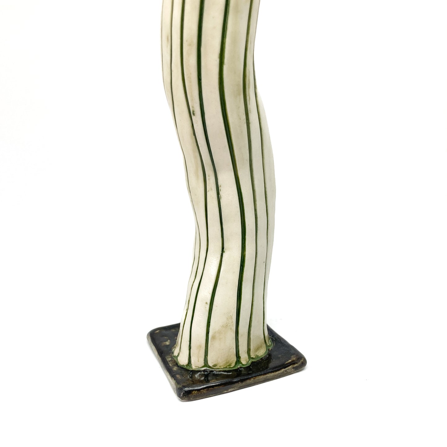 The base of a white ceramic candlestick with dark green vertical stripes and a wavy form is shown. The square base is also dark green.