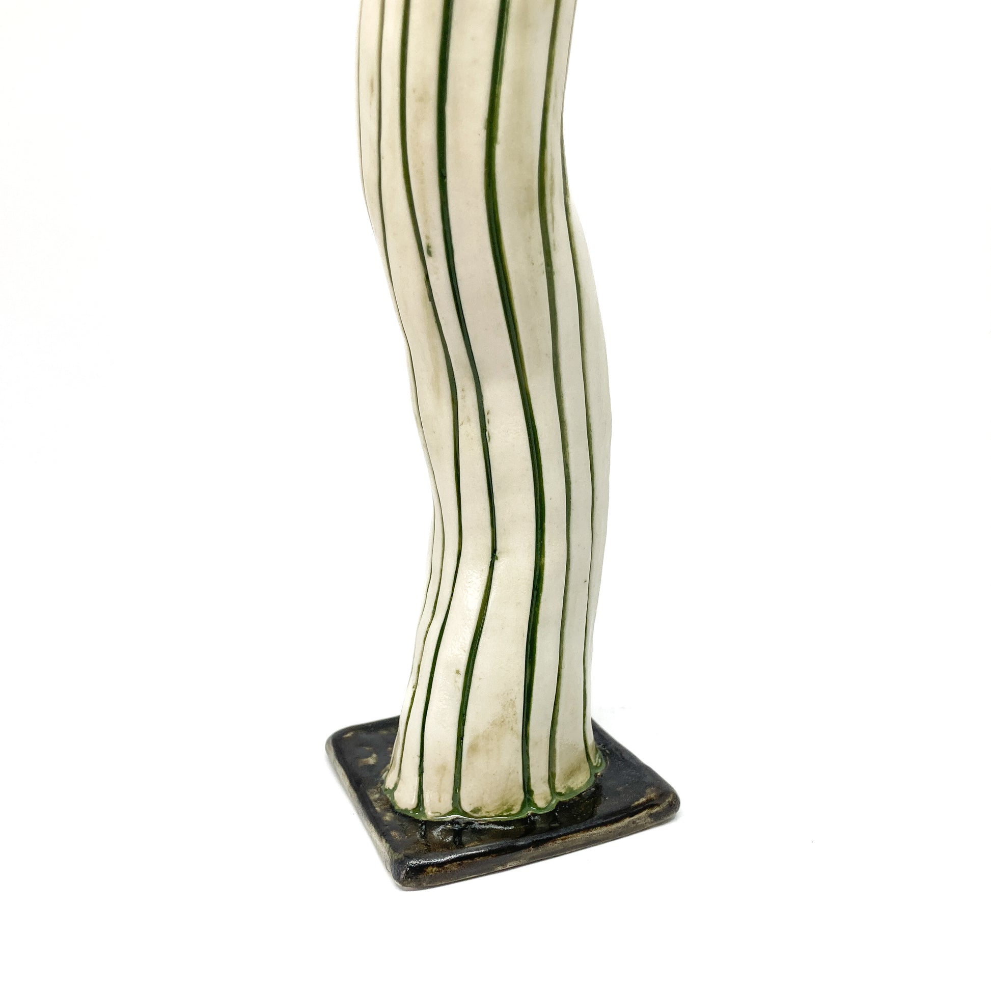 The base of a white ceramic candlestick with dark green vertical stripes and a wavy form is shown. The square base is also dark green.