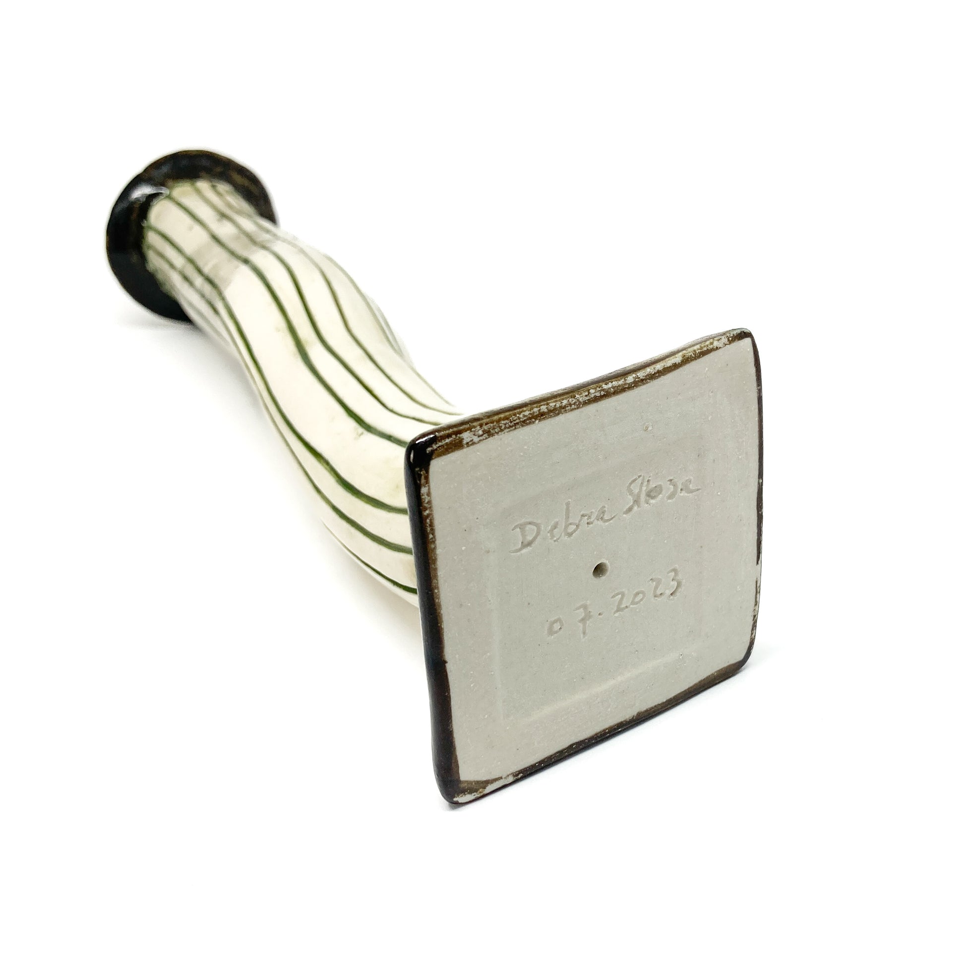 A white ceramic candlestick with dark green vertical stripes and a wavy form lies horizontally, showing its square base with an inscription "Debra Sloan 07.2023".