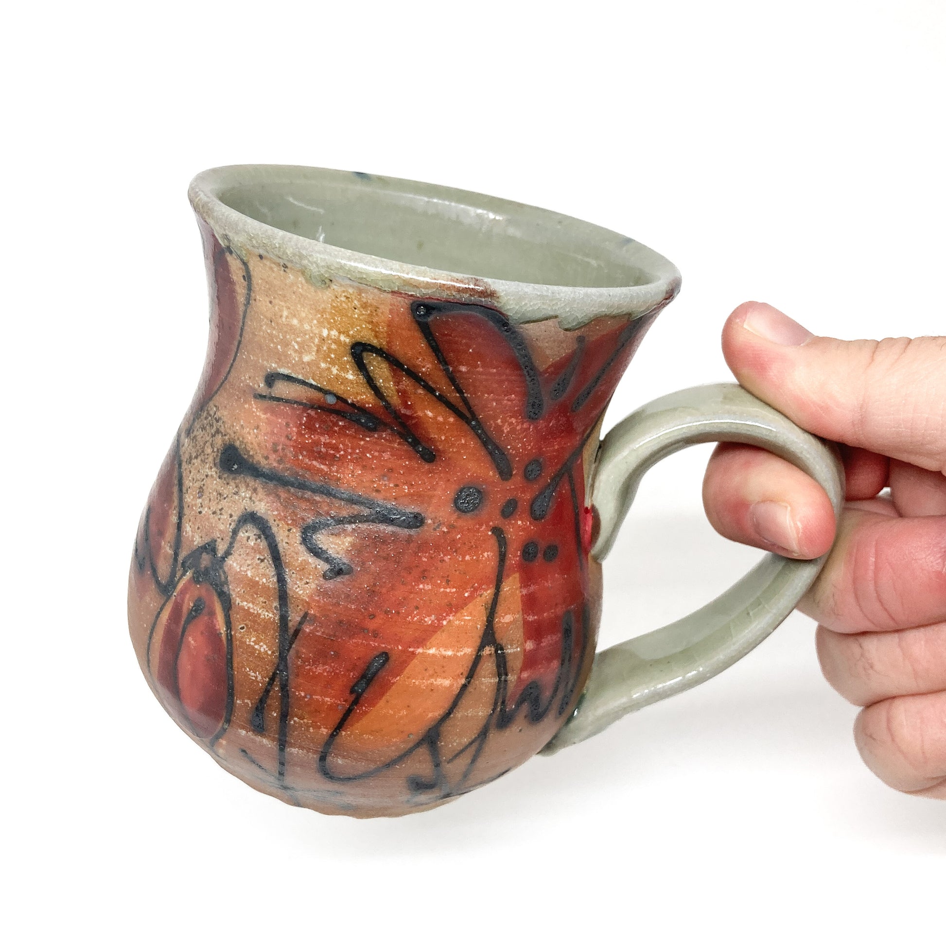 A hand holding a ceramic mug with a handle, showing a hand-painted floral design. The mug has a slightly flared shape and a smooth, glazed finish.