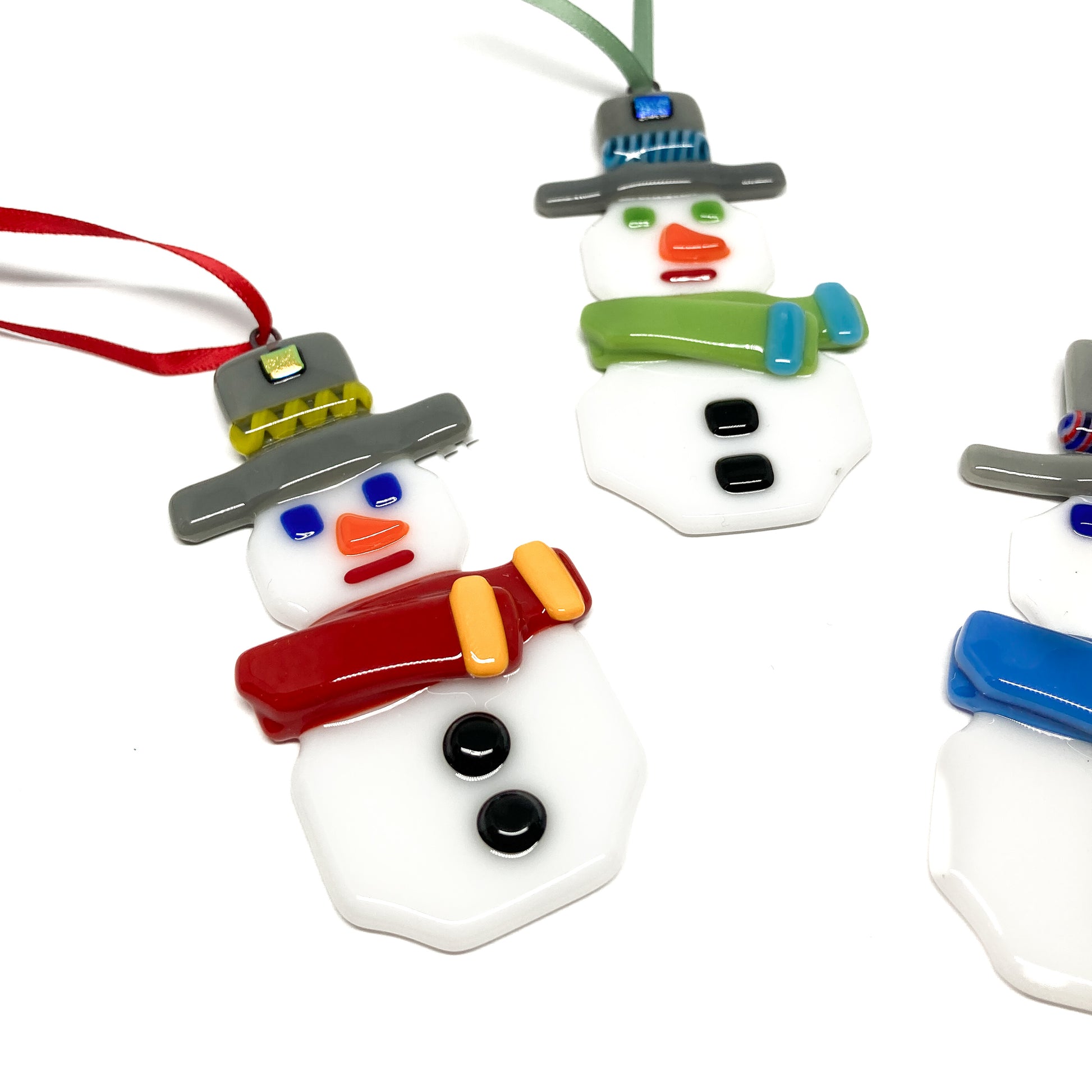 Close up of three fused glass snowman ornaments with colorful scarves and hats, each with a ribbon for hanging.