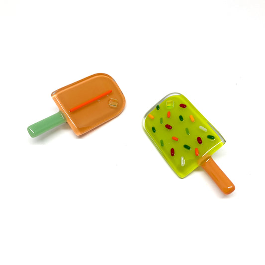 Two glass pins shaped like popsicles, one orange and one green, with colorful details.