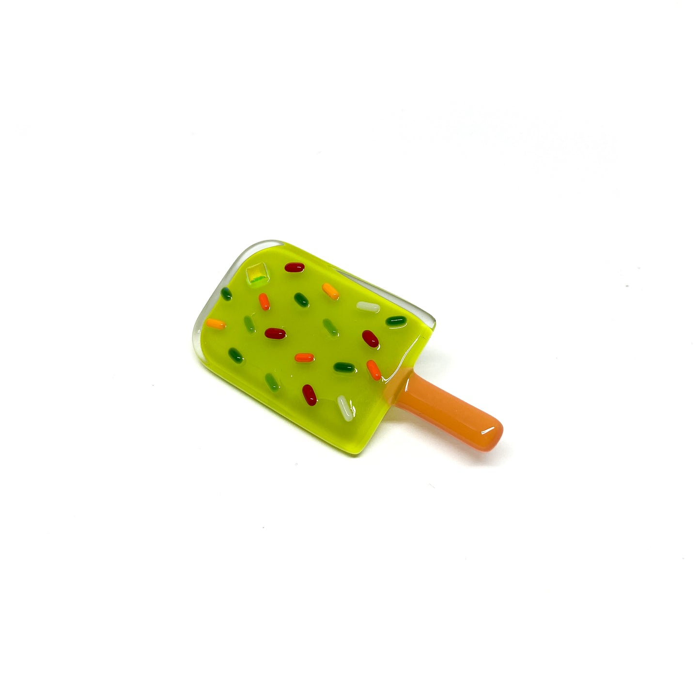 A glass pin shaped like a green popsicle with colorful sprinkles and an orange stick.