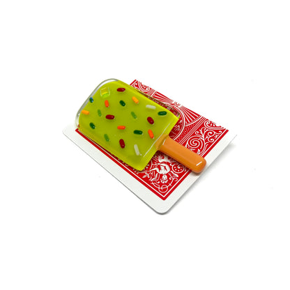 A green glass pin shaped like a popsicle with colorful sprinkles, placed on a playing card.