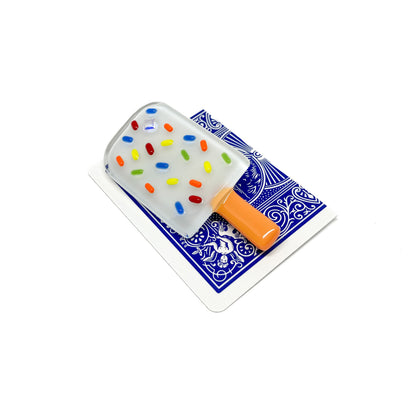 A white glass pin shaped like a popsicle with colorful sprinkles, placed on a playing card.