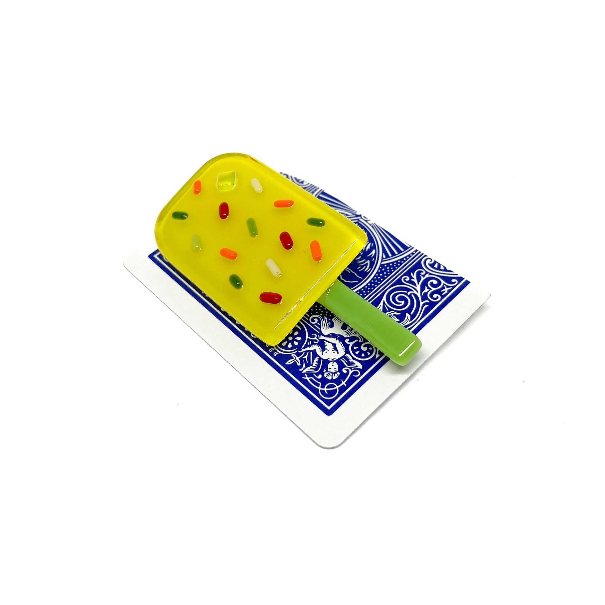 A yellow glass pin shaped like a popsicle with colorful sprinkles, placed on a playing card.