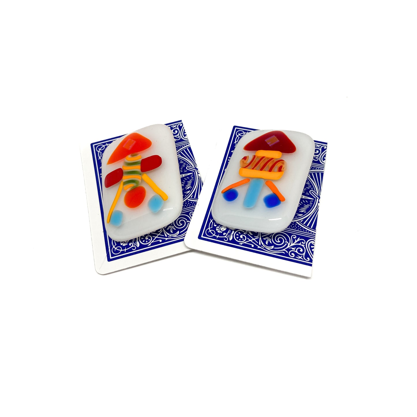 Two white glass pins with colorful abstract designs, each placed on a playing card.
