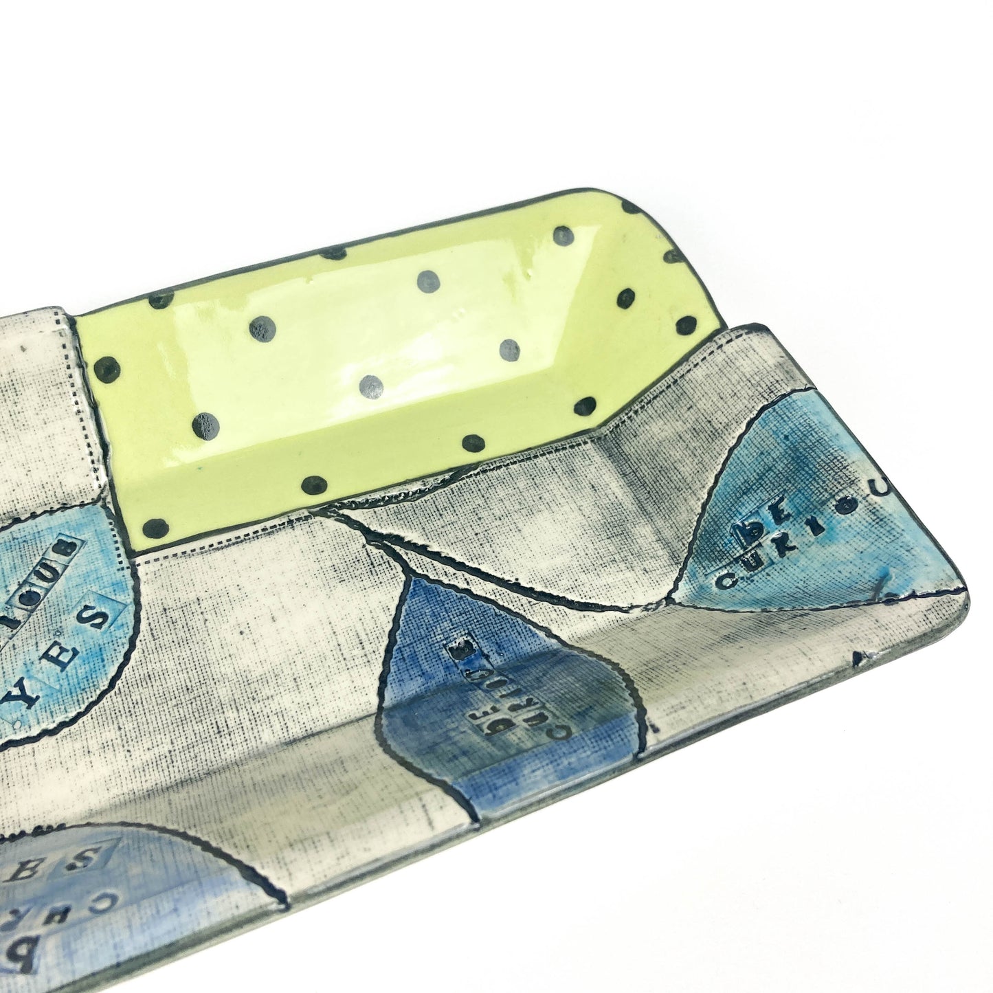 Close-up of ceramic platter corner showing polka dot glaze and hand-stamped “Be Curious” leaves.