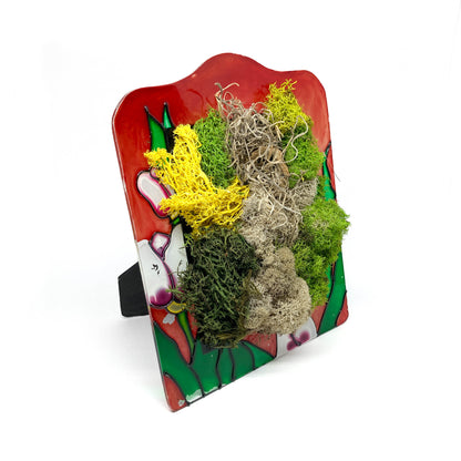 Framed artwork with preserved moss in various colors and textures, propped up on a stand, set against a background with floral designs.