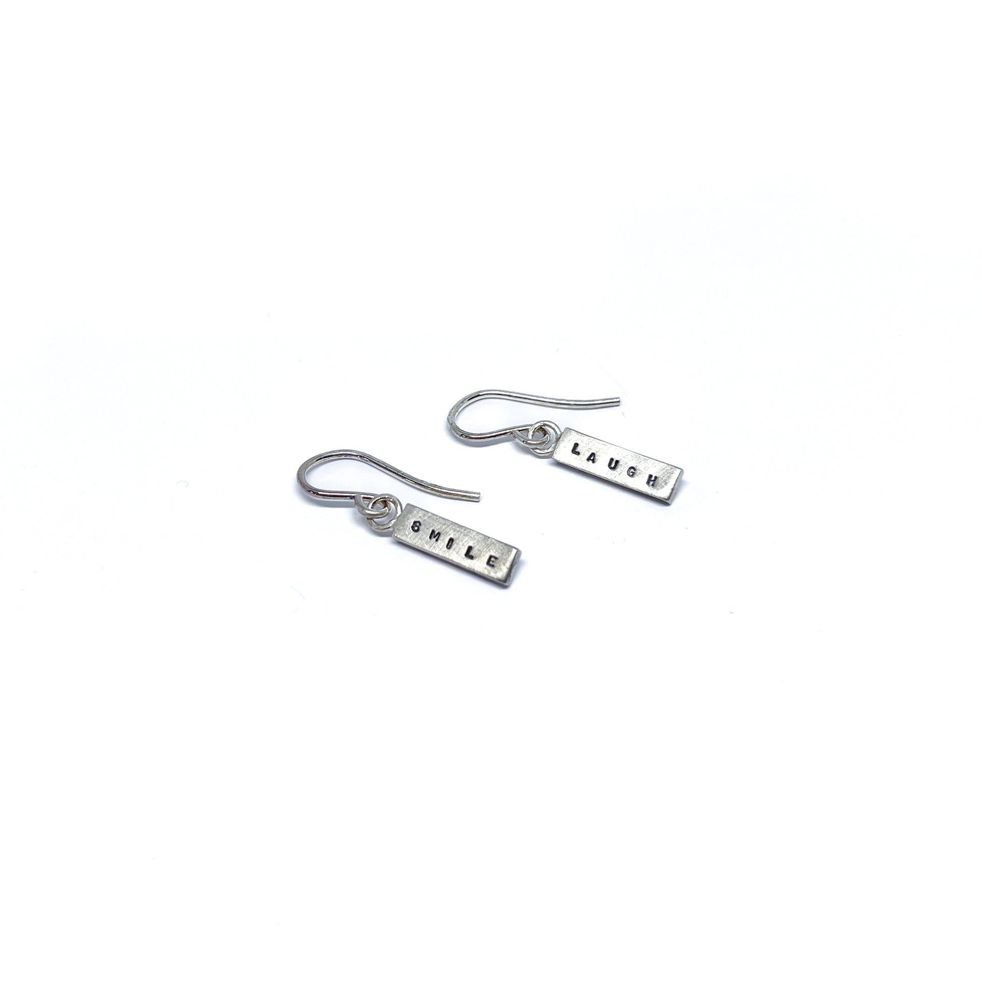 Two silver earrings with rectangular charms, one reads "SMILE" and the other "LAUGH".