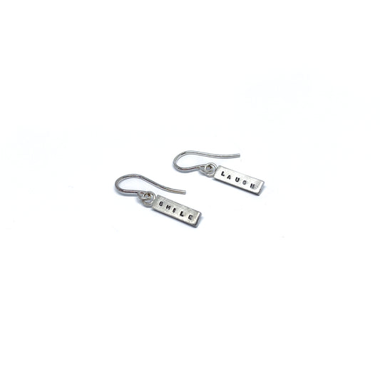 Two silver earrings with rectangular charms, one reads "SMILE" and the other "LAUGH".