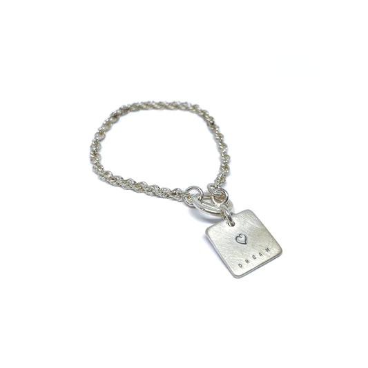 Silver bracelet with a square charm with a heart and the word "DREAM" engraved on it.