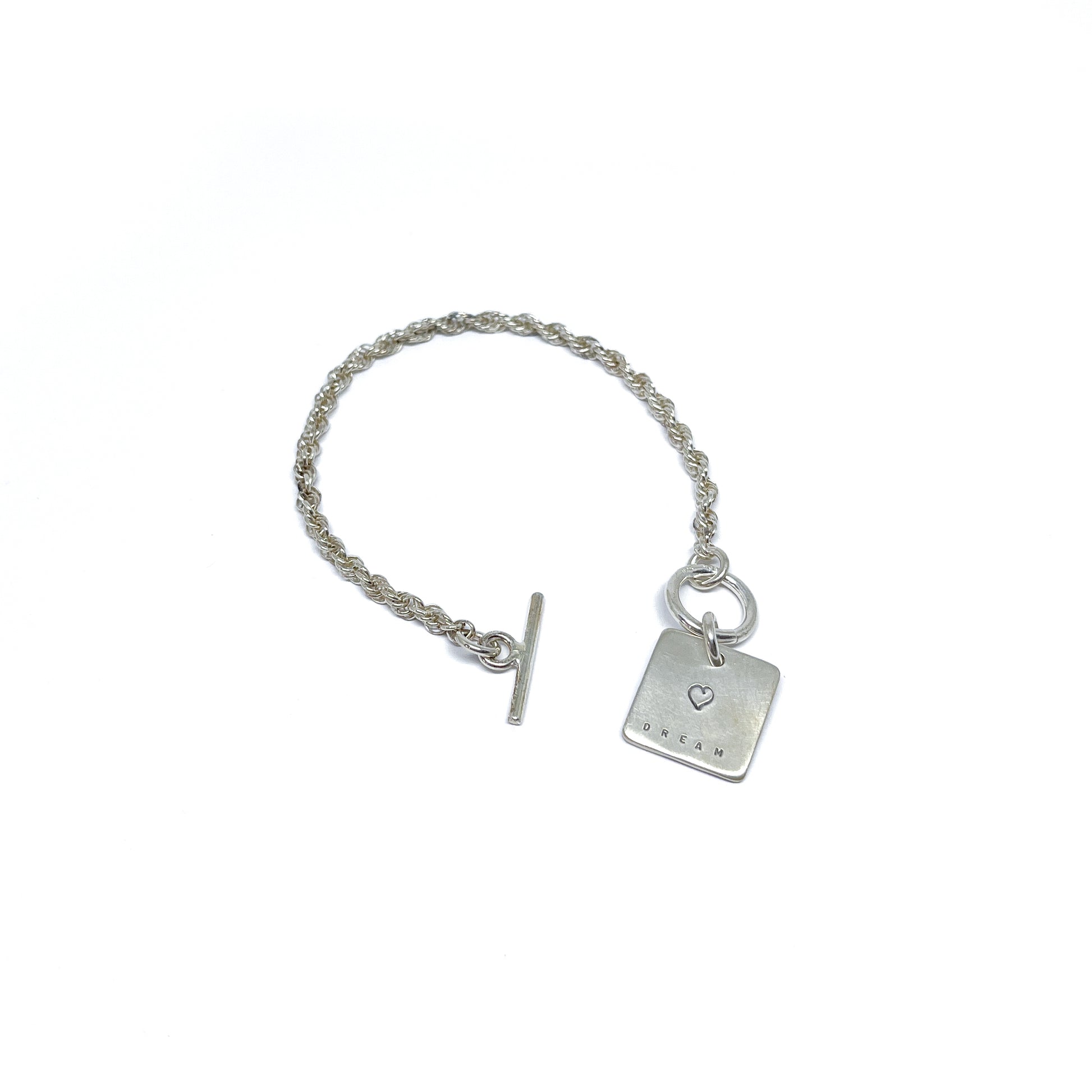 Silver bracelet with a square charm with a heart and "DREAM" engraved on it, with a t-bar clasp.