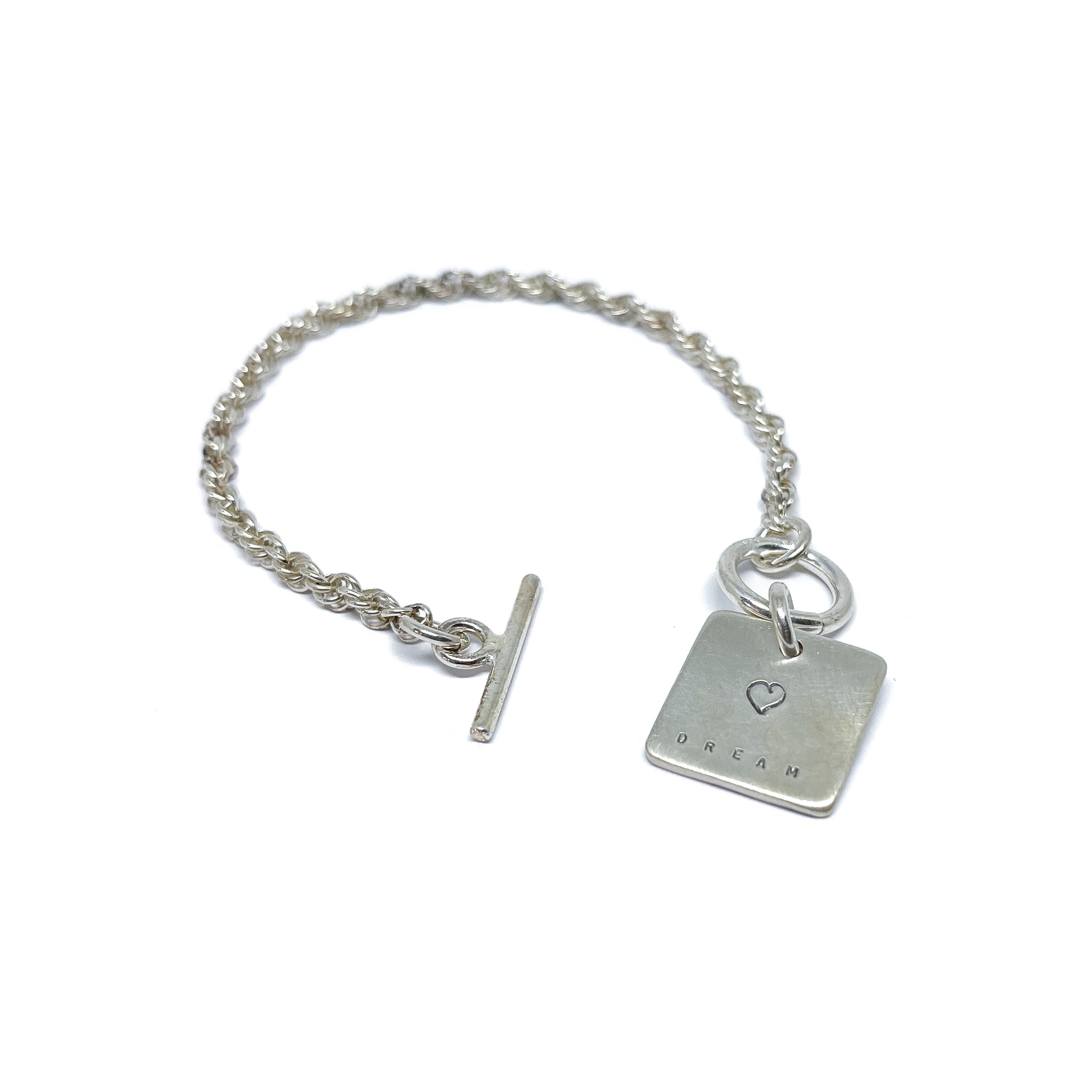 Silver bracelet with a square charm with a heart and "DREAM" engraved on it, with a t-bar clasp.