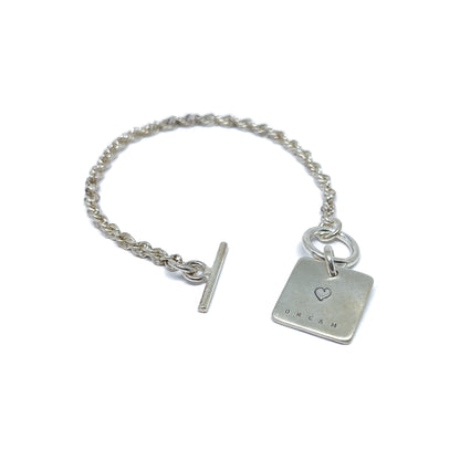 Silver bracelet with a square charm with a heart and "DREAM" engraved on it, with a t-bar clasp.