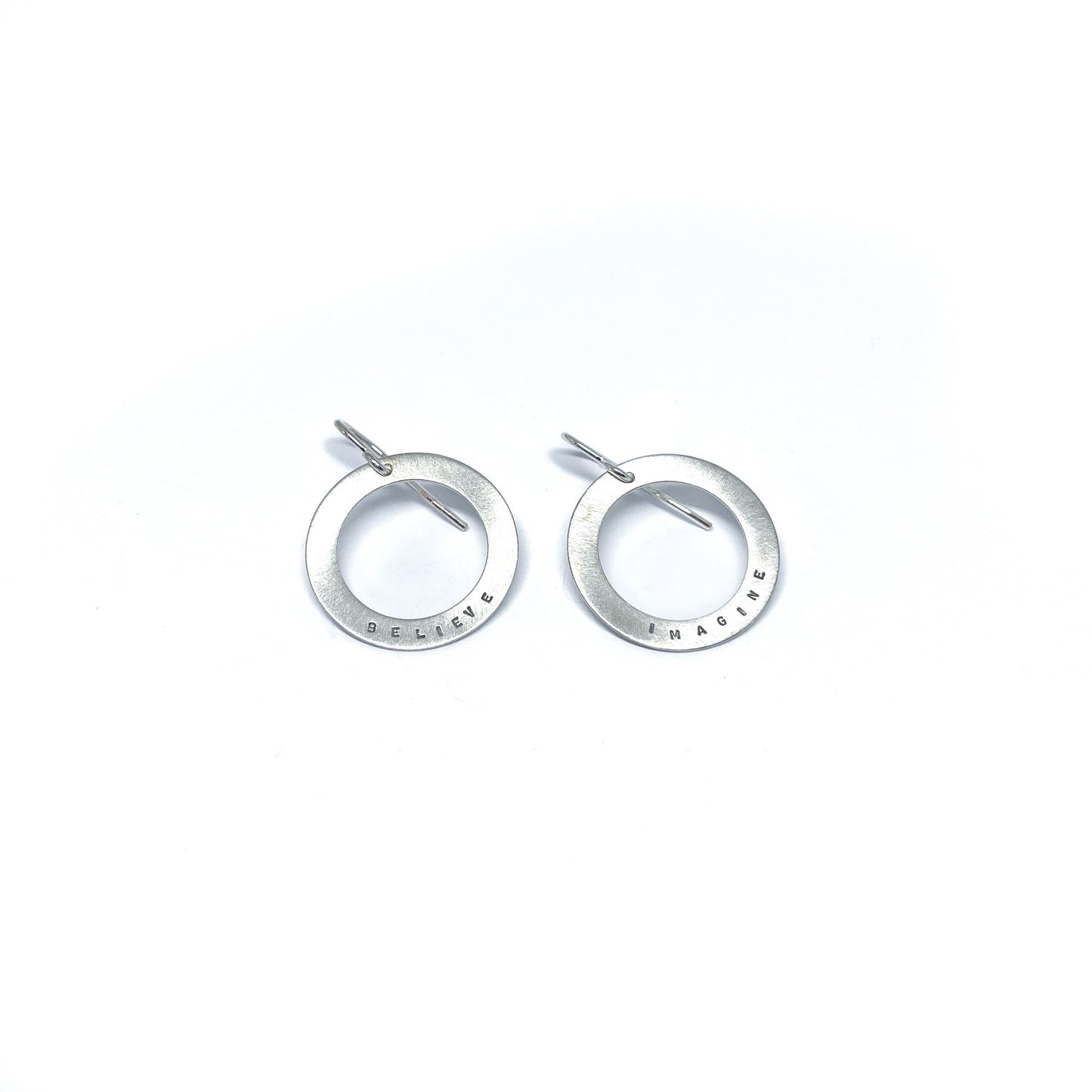 Two silver earrings with circular pendants, one engraved with "BELIEVE" and the other with "IMAGINE."