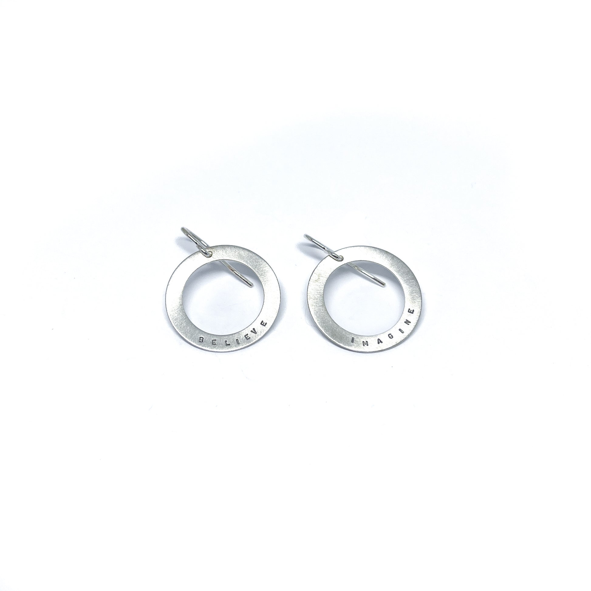 Two silver earrings with circular pendants, one engraved with "BELIEVE" and the other with "IMAGINE."
