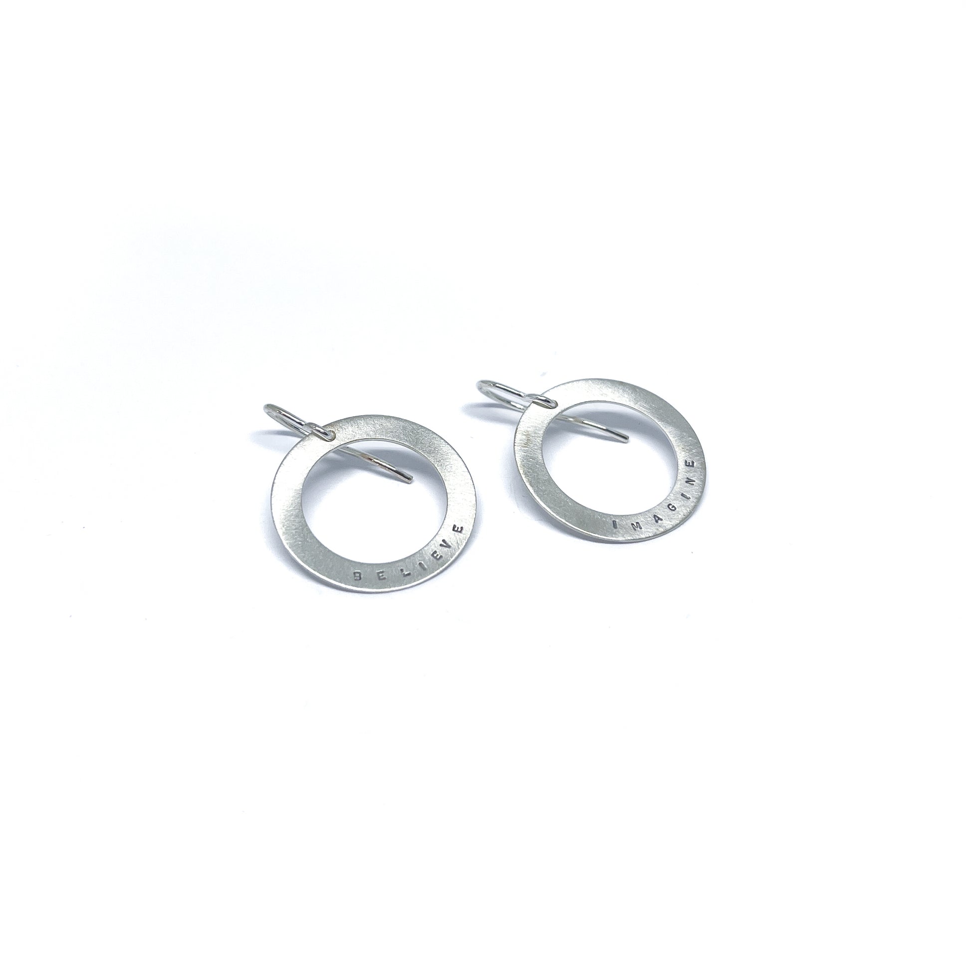 Two silver earrings with circular pendants, one engraved with "BELIEVE" and the other with "IMAGINE."